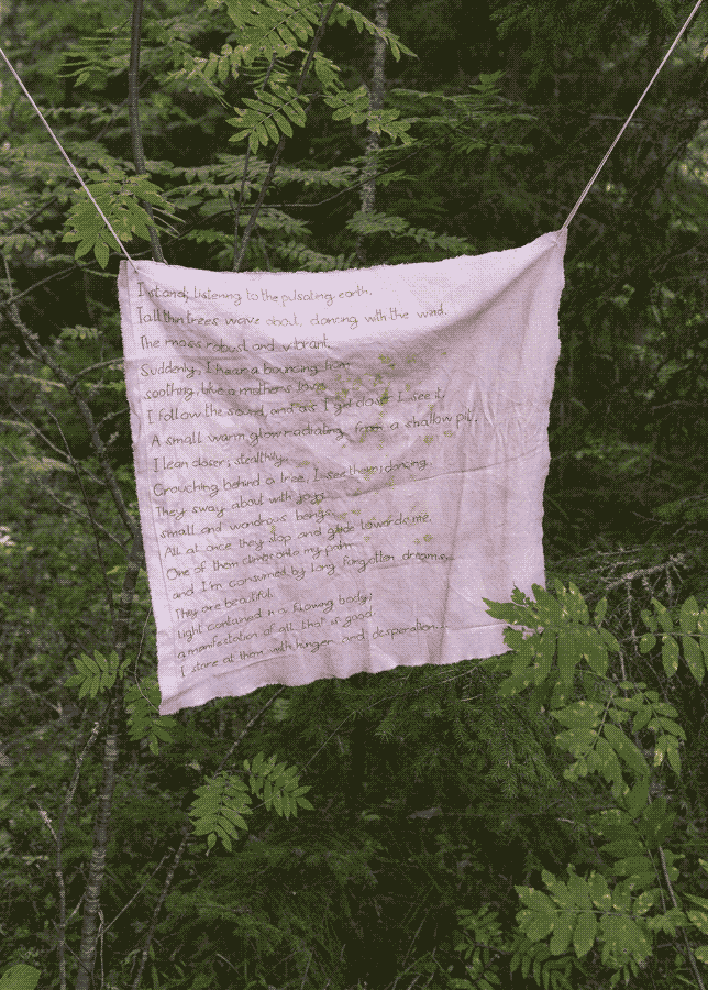 Close up of the second fabric hanging in the forest. It is light pink and contains part of a stitched poem.