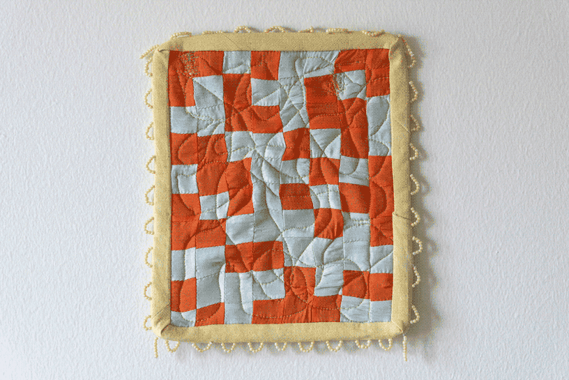 A small and square quilt made of orange and light blue checkers.
                    The binding is a pale yellow and the border is decorated with white white beaded loops.