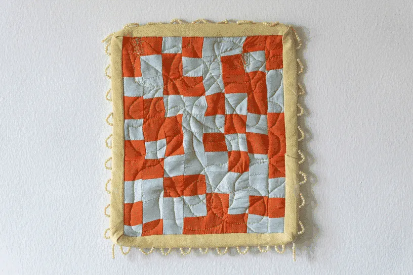 A small and square quilt made of orange and light blue checkers.
                            The binding is a pale yellow and the border is decorated with white white beaded loops.