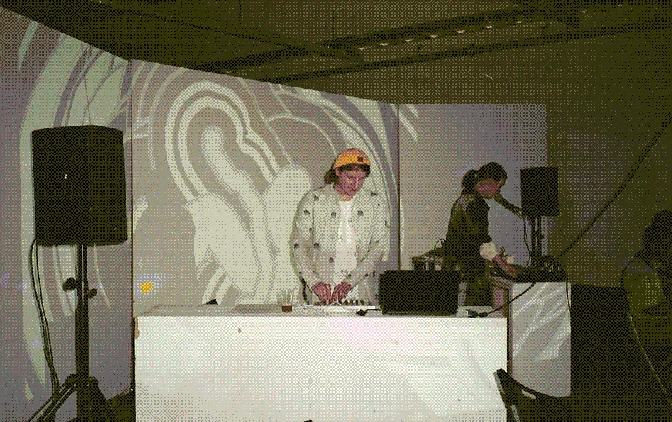 Marloes van Son and Alex stand behind a platfrom topped with music equipment. 
                    Large speakers sit at each end of the platform.