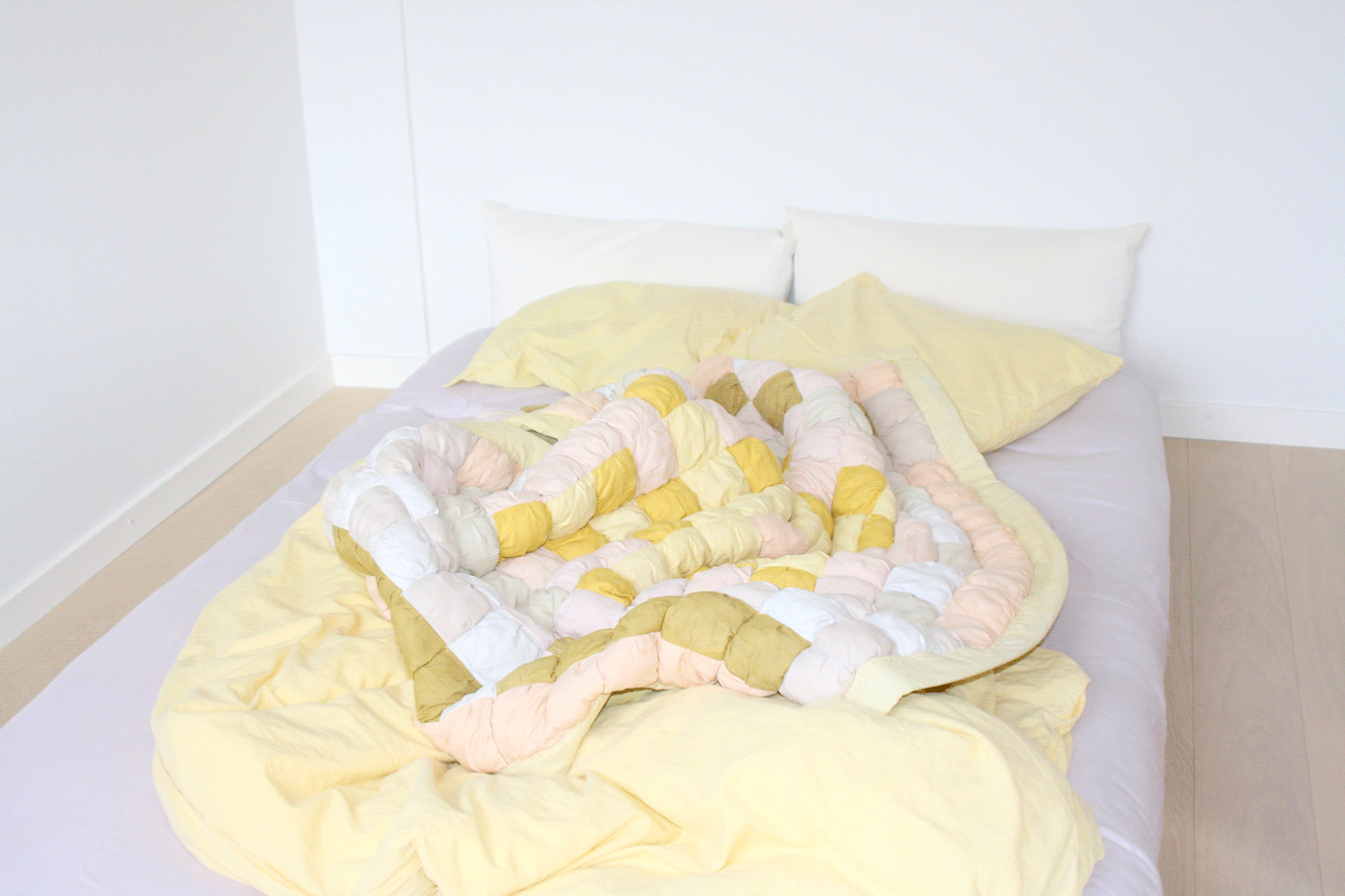 A puffy quilt lays on a messy bed. The quilt has pale tones
                of yellows, greens, and pinks.