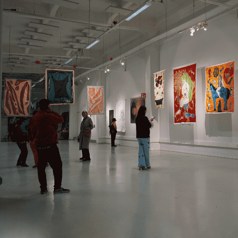 A white hall filled with an arrangement of hanging textile artworks.
                        on the wall there are paintings and drawings. Scattered thoughout the
                        space are visitors examining the artworks.