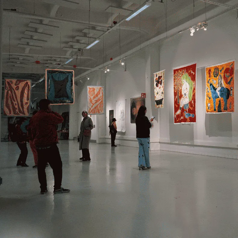 A white hall filled with an arrangement of hanging textile artworks.
                            on the wall there are paintings and drawings. Scattered thoughout the
                            space are visitors examining the artworks.