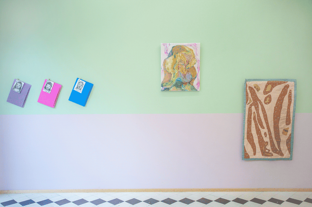 A green and pink wall of an exhibition space. From left to right on the wall, there are:
                small ink drawings attached to office envelopes, a medium size yellow painting of a human-like
                figure, and a brown quilt with organic shapes.