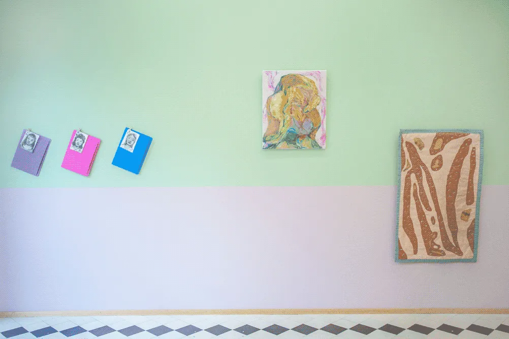 A green and pink wall of an exhibition space. From left to right on the wall, there are:
                    small ink drawings attached to office envelopes, a medium size yellow painting of a human-like
                    figure, and a brown quilt with organic shapes.