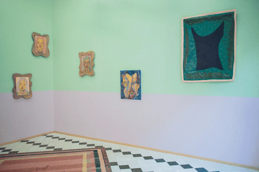 the same exhibition space, but from a different angle. From left to right, on 
                    the wall there are: three small paintings of human-like figures in wooden frames placed in
                    a dyniamic arrangement on the wall, a medium sized blue panting of a human-like figure, 
                    and a large green quilt with abstract and organic shapes. There is also a large red rug 
                    at the center of the room.