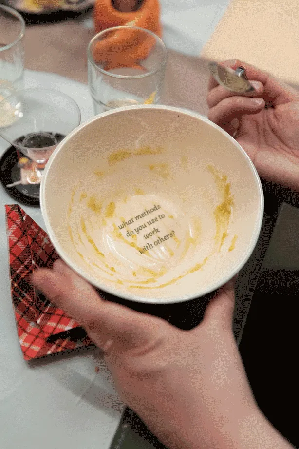 A close up of one hand hondling a soup bowl, while the other hand, holds the spoon.
                            Both hands are leaning against the table, and on the table are various glasses and a napkin.
                            Inside the bowl it reads what methods do you use to work with others?.