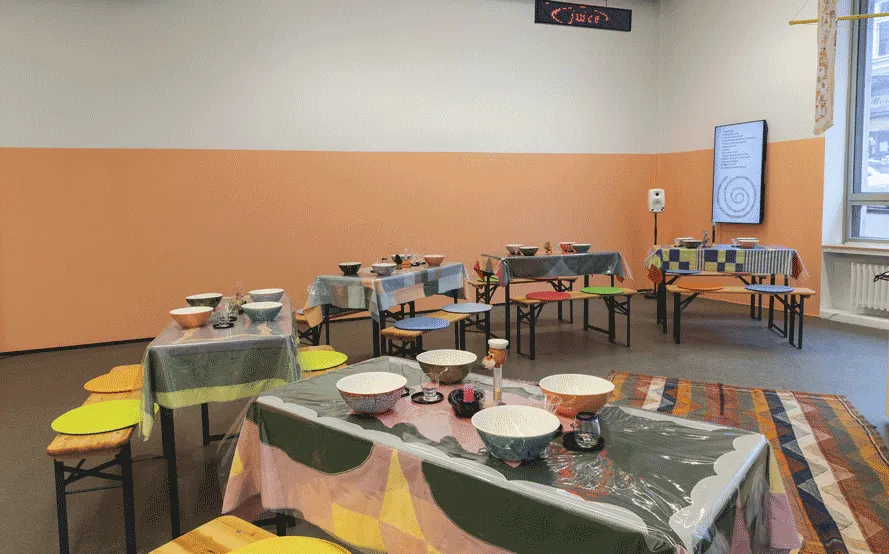 The viewer is facing a corner of the gallery. Botton half of the wall is painted a coral pink.
                    At the very corner of the space there is a tv mounted on the wall with the exhibition's visuals and related 
                    content. Hanging from the celiing is an lcd sign flashing soup ingredients in red pixel text. In the room 
                    there there five tables forming a semi-circle with a large woven carpet in the center. The tables all have 
                    different and colorful tablecloths. The tables are set with cutlery and dishware. The benches that accompany 
                    the tables have colorful circular cushions.