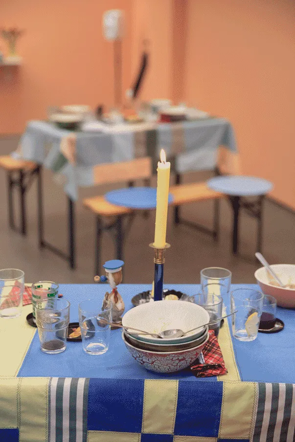 A closeup of a dining table. The table is covered by a handmade tablecloth made by 
                            the artist with rectangular shapes varying in size and colors of blue, yellow and
                            green and white strips. In the center there is a candle holder with a lit candle, and 
                            around it, are empty glasses, a stack of soup bowls with a few spots in the topmost bowl, and 
                            tiny black trays scattered on the table. The are a few red napkinds and a tiny table sculpture.