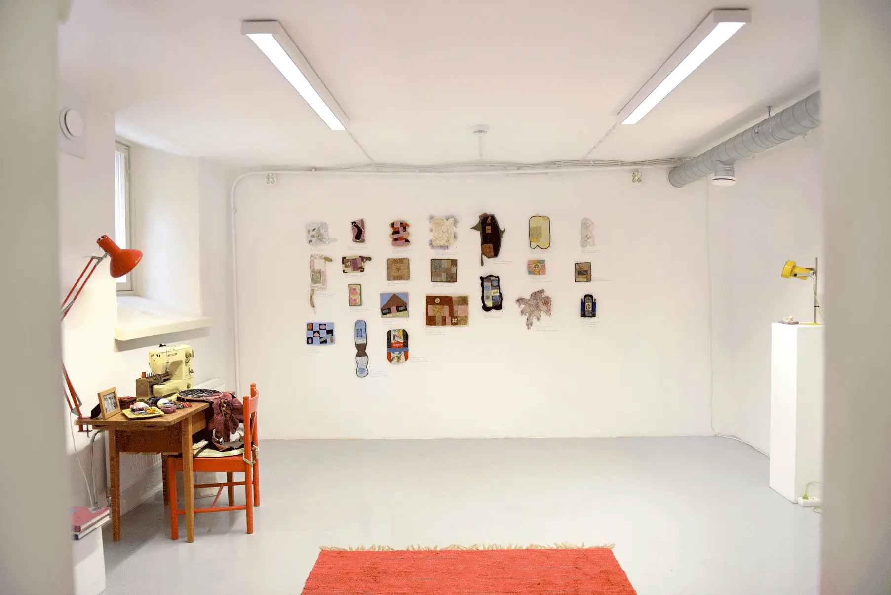 The viewer is facing the exhibition space from the entry point. 
                    There is a red carpet on the floor. To the left of the room is a sewing table and on it is an 
                    old sewing maching, sewing materials, a red lap, a picture frame of the artist's family 
                    and orange chair. To the right is a podium and a yellow table lap sets on it.
                    and in front of the viewer are many small quilts in various shapes, colors and sizes, but 
                    all are relatively small.