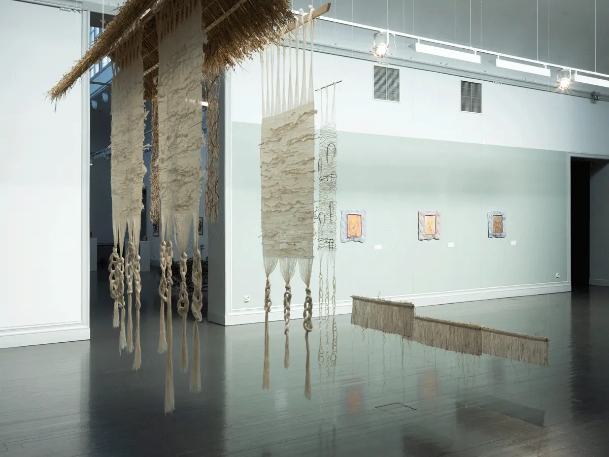 the viewer looking from the corner of the gallery. Right in front are several woven pieces hanging from the ceiling. 
                    and on the wall, are the three paintings resembling portraits of three different women.