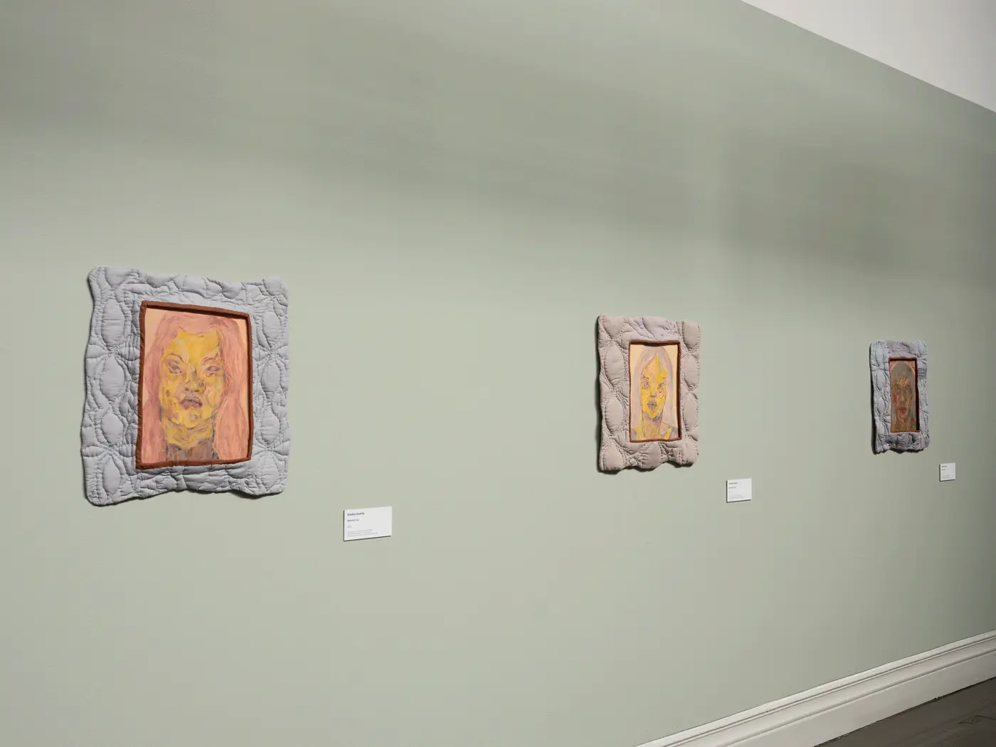 The viewer is facing a pale green wall, and on the wall, are three, evenly spaced pantings. 
                    each painting has a bluegrey quilted frame. The paintings are resemble portraits of women.