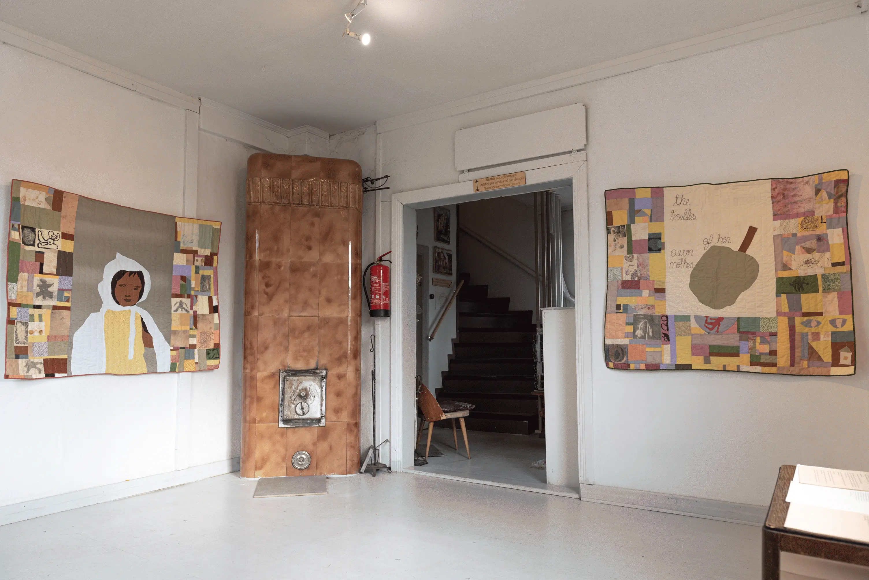 The viewer is facing the exhibition space from the corner of the room. 
                    To the left of the room, on the wall is a quilt with two three panels. The outer panels are of colorful patchwok,
                    and the middle panel is a portrain of a child in a white cape. in the middle is a doorway to another room, and
                    to the right is another wall with a quilt similar to the precious one, but the central panel has a fruit and a text
                    that reads: The troubles of her own mother.