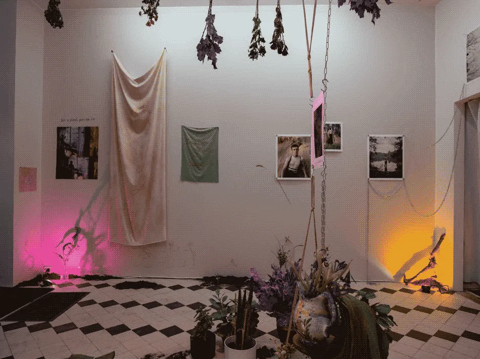 The same space, but from another angle. On the wall hangs textile works and more photographs of
                    bodies and nature. There are small piles of dirt around the space with pink and orange spotlights at the
                    corners of the walls.
