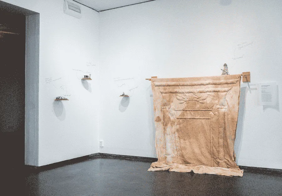 The same gallery space but from a different angle. There are several small 
                    ceramic figurines places on little shelves on the wall. They have a dynamic arrangement
                    that lead to hanging mantle made of textile.